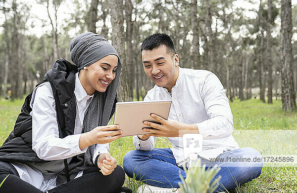 Lächelnder junger Mann und schöne Frau benutzen ein digitales Tablet  während sie im Wald sitzen