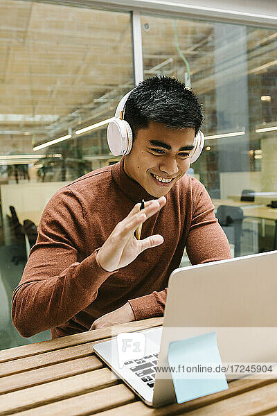 Lächelnder männlicher Unternehmer  der während eines Videogesprächs am Laptop in der Cafeteria mit der Hand winkt