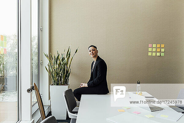 Lächelnde Geschäftsfrau sitzt auf dem Schreibtisch an der Wand im Büro