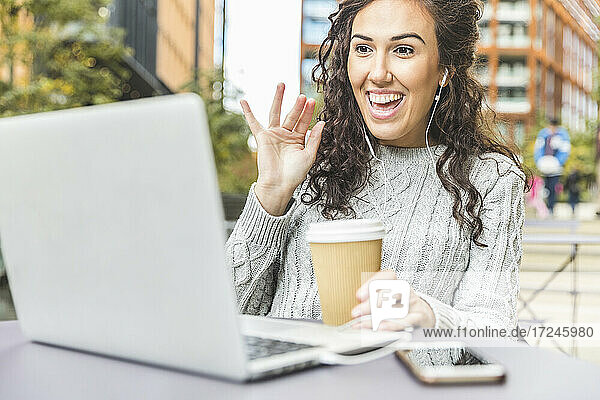 Lächelnde weibliche Fachkraft winkt während eines Videoanrufs über einen Laptop in einem Café