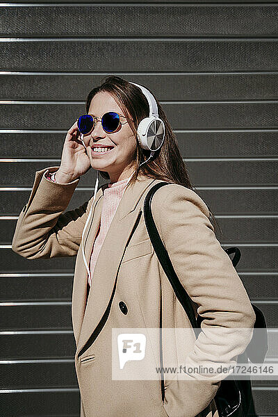 Junge Frau mit Kopfhörern und Sonnenbrille an der Wand stehend