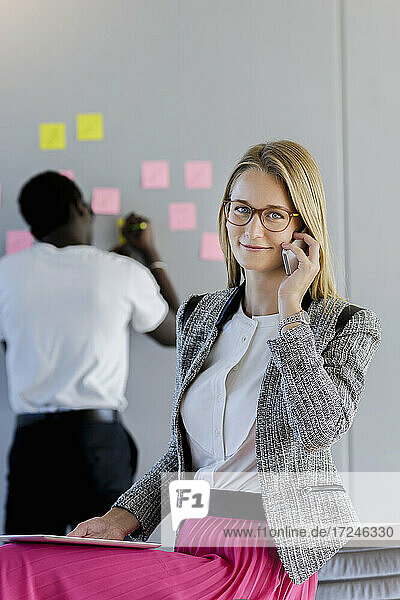 Weibliche Fachkraft  die mit einem Smartphone spricht  während ein männlicher Kollege im Hintergrund im Büro sitzt