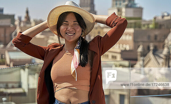 Lächelnde Frau mit Mütze steht mit den Händen hinter dem Kopf auf einer Dachterrasse