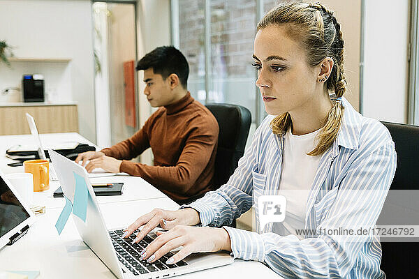 Junge Geschäftsfrau  die einen Laptop benutzt  während sie mit einem Kollegen im Hintergrund im Büro sitzt