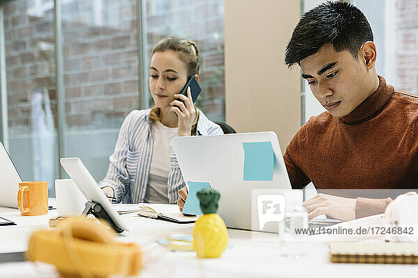 Geschäftsmann  der an einem Laptop arbeitet  neben einer Kollegin  die über ihr Smartphone in einem Coworking-Büro spricht