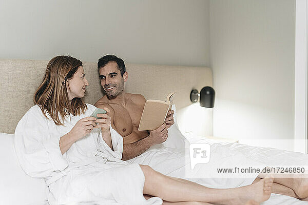 Mann ohne Hemd mit Buch schaut Frau an  während er auf dem Bett im Hotelzimmer sitzt