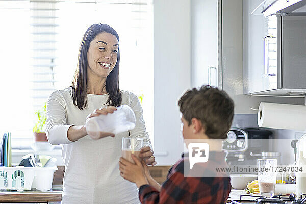 Lächelnde Frau gießt Milchshake in ein Glas  das ihr Sohn in der Küche hält