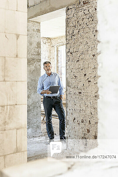 Männlicher Architekt mit Hand in der Tasche  der ein digitales Tablet auf einer Baustelle hält