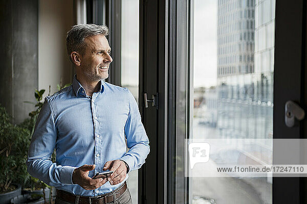 Smiling businessman looking through window while holding mobile phone at work place