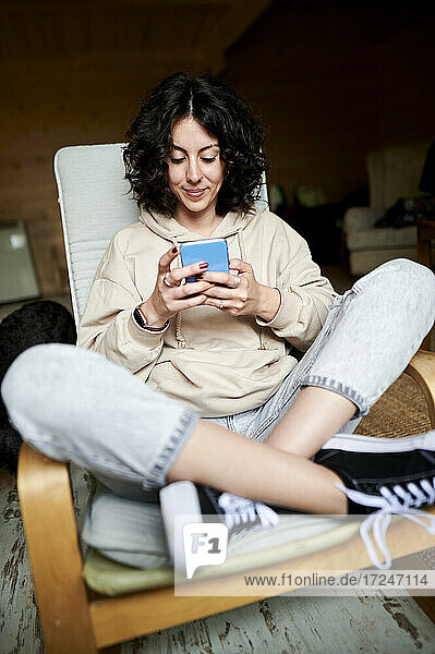 Lächelnde Frau sitzt mit gekreuzten Beinen und benutzt ein Mobiltelefon zu Hause