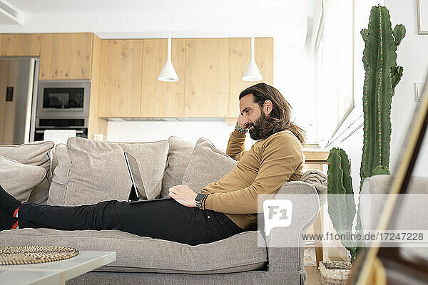 Mann mit langen Haaren  der einen Tablet-PC benutzt  während er sich auf dem Sofa zu Hause entspannt