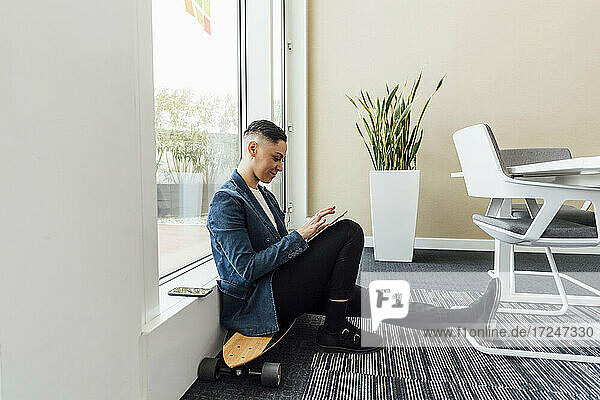 Unternehmer  der ein digitales Tablet benutzt  während er im Büro auf einem Skateboard sitzt