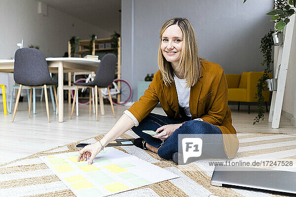 Smiling creative businesswoman with adhesive notes sitting on carpet in office