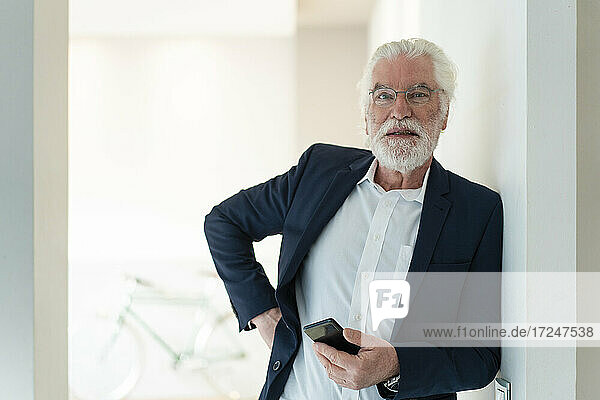 Älterer Geschäftsmann mit Smartphone an der Wand lehnend im Büro