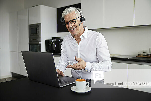 Lächelnder Geschäftsmann mit drahtlosem Headset  der über einen Laptop in der Cafeteria ein Videogespräch führt
