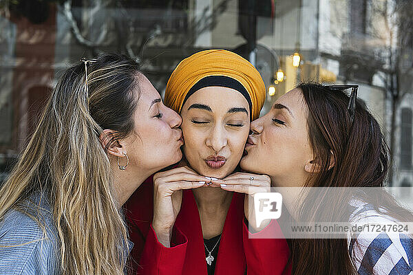 Female friends kissing young woman wearing headscarf outdoors