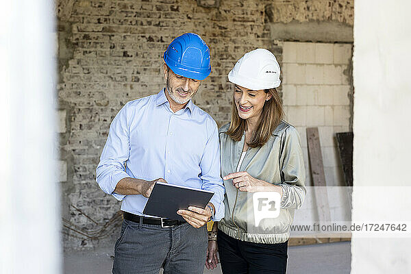 Weiblicher Bauherr und männlicher Architekt mit Schutzhelmen diskutieren auf einer Baustelle über ein digitales Tablet