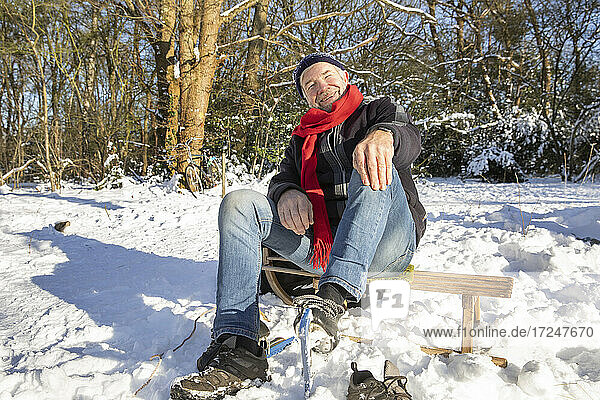 Glücklicher älterer Mann  der an einem sonnigen Tag auf einem Schlitten im Schnee sitzt