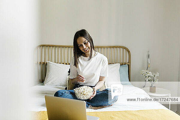 Lächelnde junge Frau  die Popcorn isst und auf einen Laptop auf dem Bett schaut