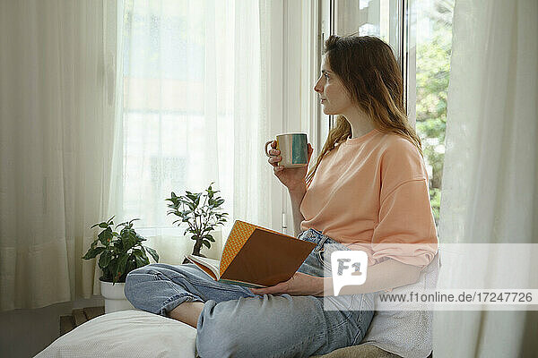 Nachdenkliche Frau mit Buch  die wegschaut  während sie zu Hause auf einer Matratze sitzt
