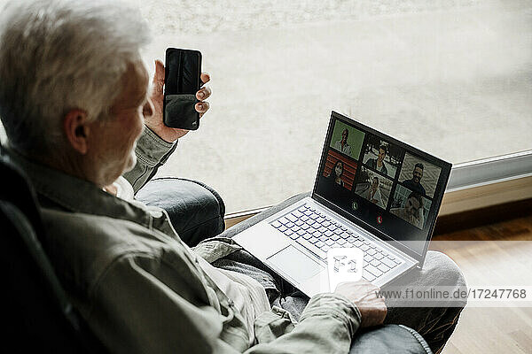 Senior man with mobile phone talking on video call through laptop at home