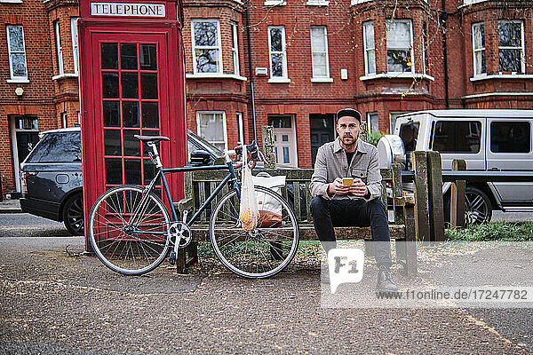 Man with smart phone sitting on bench near telephone booth at street