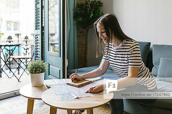 Lächelnde Frau schreibt eine Notiz in ihr Tagebuch  während sie am Tisch in einer Wohnung sitzt