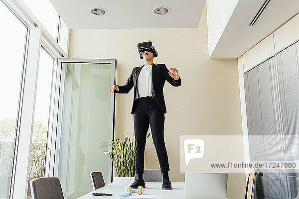 Young businesswoman using virtual reality simulator while standing on desk in office