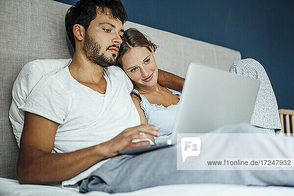 Young man using laptop while sitting by woman in bed at home