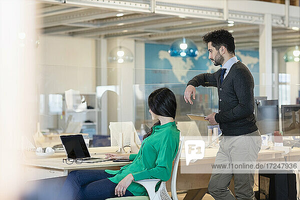 Young businessman with digital tablet standing behind female colleague using laptop in office