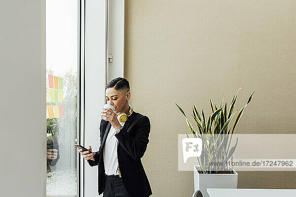 Junger Unternehmer trinkt Kaffee  während er im Büro sein Mobiltelefon benutzt
