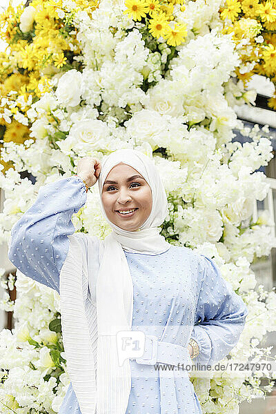 Lächelnde Frau mit Hand auf der Hüfte vor weißen Blumen stehend
