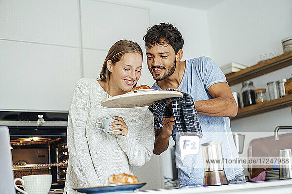 Man showing freshly baked croissant to girlfriend in kitchen at home