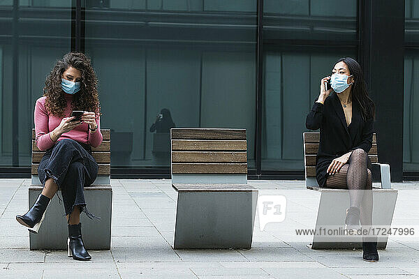 Frau mit Gesichtsmaske  die ein Mobiltelefon benutzt  während sie in sozialer Distanz mit einer Frau auf dem Gehweg sitzt