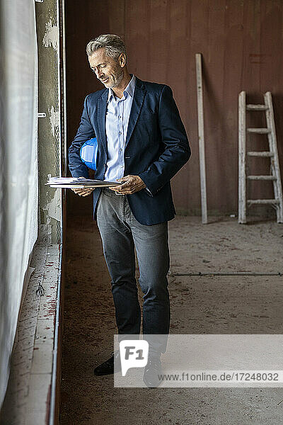 Männlicher Architekt sieht sich Dokumente an  während er auf der Baustelle am Fenster steht