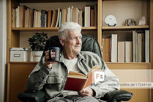 Senior man with book looking away while holding mug at home