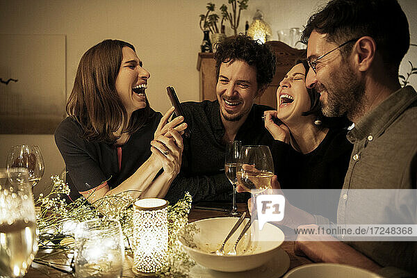 Männliche und weibliche Freunde lachen  während die Frau am Esstisch ihr Handy zeigt