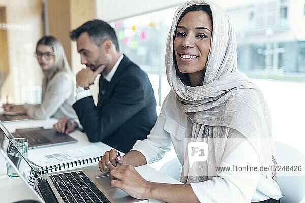 Lächelnde Geschäftsfrau mit Kopftuch am Laptop im Büro