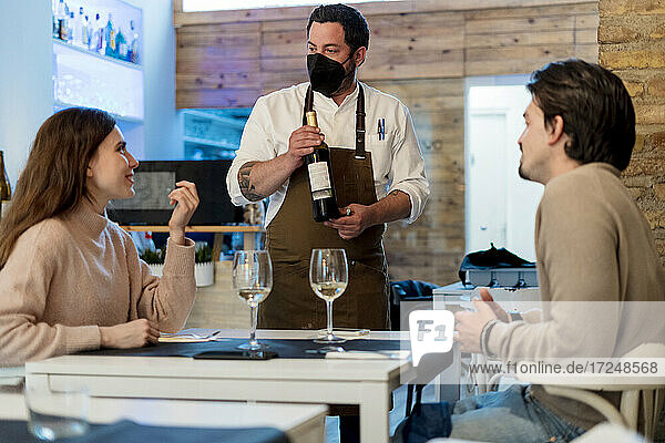 Ehepaar im Gespräch mit dem Kellner  der eine Weinflasche im Restaurant hält  während COVID-19