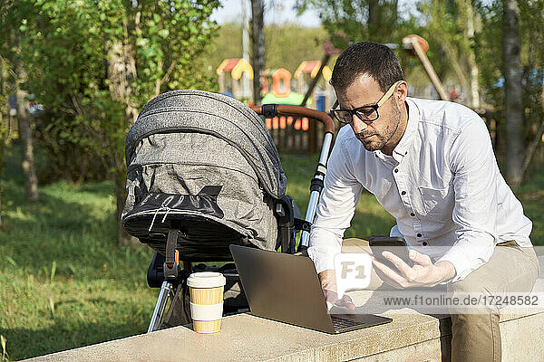 Male entrepreneur holding mobile phone while using laptop by baby stroller in park