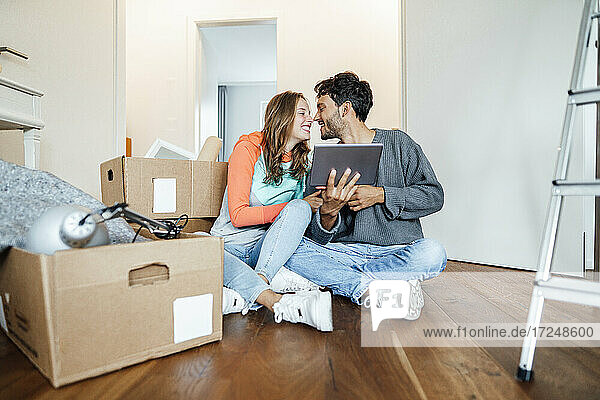 Glückliches junges Paar mit Tablet  das neben Kartons im neuen Haus sitzt