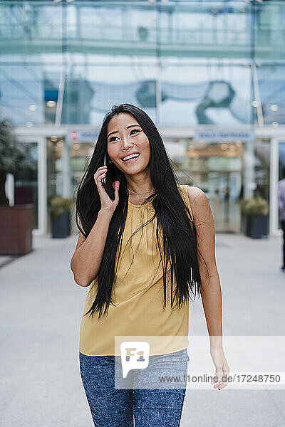 Lächelnde schöne Frau  die am Telefon spricht  während sie vor einem Bürogebäude geht