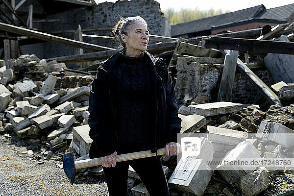 Mature woman in jacket holding hammer while standing by demolished house
