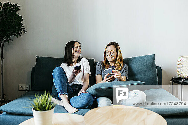 Lächelnde Freundinnen  die zu Hause auf dem Sofa sitzen und ein Mobiltelefon benutzen