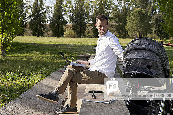 Father looking at baby stroller while sitting with laptop at park