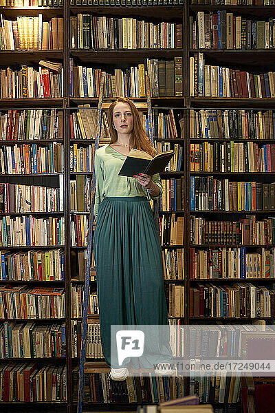 Junge Frau hält ein Buch und steht auf einer Leiter in einer Bibliothek