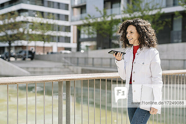 Fröhliche Frau  die eine Voicemail über ihr Smartphone sendet  während sie an einem Geländer in der Stadt spazieren geht