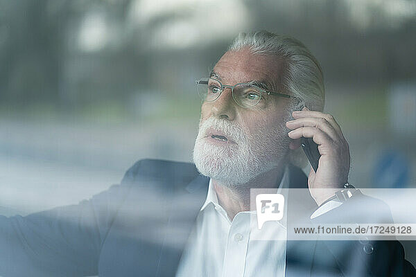 Überraschender älterer Geschäftsmann  der im Büro mit seinem Smartphone telefoniert  gesehen durch Glas