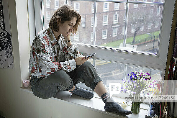 Frau benutzt Mobiltelefon  während sie zu Hause auf der Fensterbank sitzt