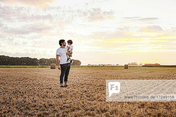 Father carrying baby son through harvested field at sunset
